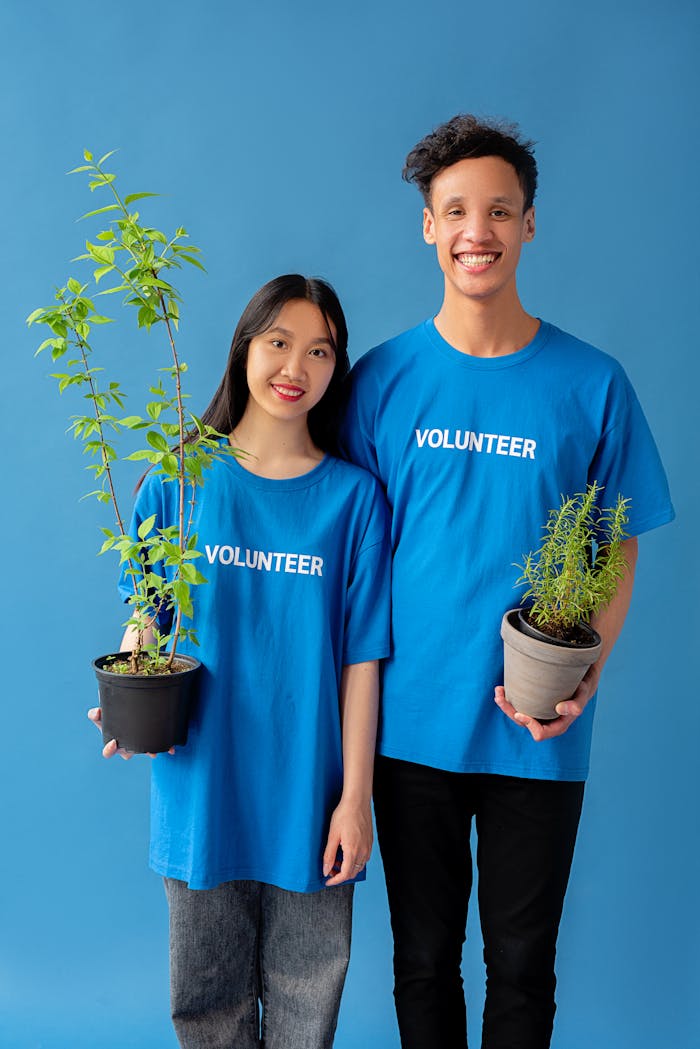 Two smiling volunteers in blue shirts hold potted plants against a blue background.